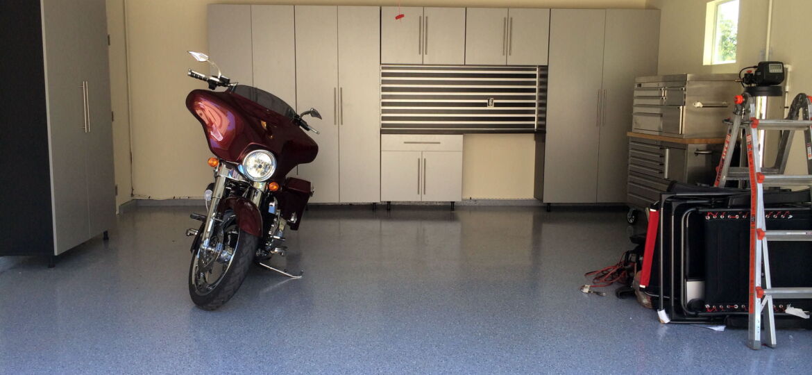 A well-organized garage with a glossy blue-speckled floor. Centered is a maroon motorcycle parked facing the garage door. To the left, a large black cabinet matches the color of the bike. On the right, a wall-mounted tool cabinet, a tall storage unit, and a workbench with multiple drawers are neatly arranged. A red fire extinguisher is mounted on the wall, and natural light streams in from a small window.