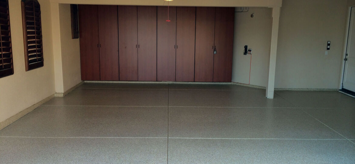 A tidy residential garage featuring a closed wooden double door, with a smooth, speckled epoxy floor. To the left, there are two windows with closed brown shutters, and to the right, a white side door with an electrical outlet and a hose bib. An opened garage door is visible at the top of the image, and a single pendant light hangs from the ceiling.