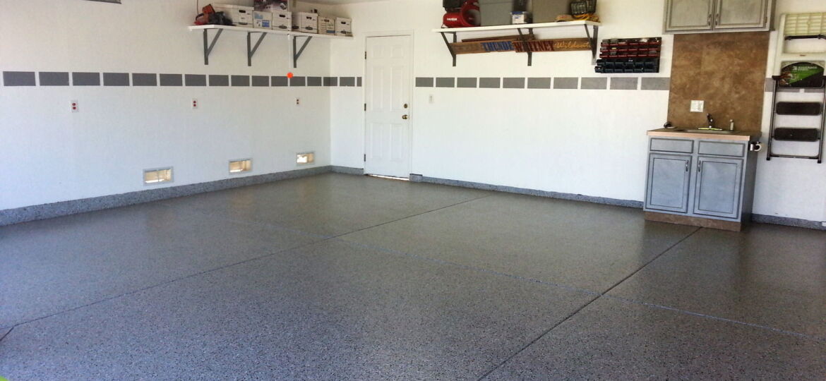 A tidy garage interior with a textured grey terrazzo floor. The walls are adorned with a horizontal stripe and have several mounted shelves holding various items, including storage boxes and a toolbox. A workbench area with a cabinet and a countertop is situated on the right side, next to a white door. The garage door's mechanical components are visible overhead. Epoxy flooring is always a great choice for garages and more!