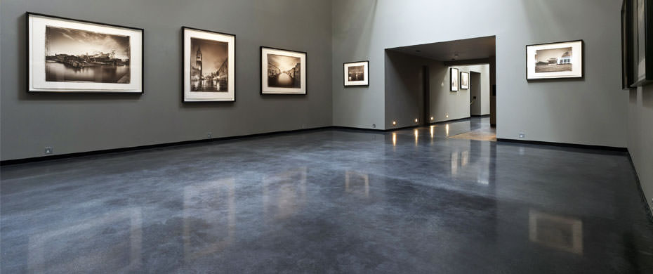 A modern art gallery with polished concrete floors in a gradient of gray tones, leading to a well-lit hallway. The left wall displays a series of framed black and white photographs of architectural landscapes, spaced evenly. The room's subdued lighting focuses attention on the artworks, creating an atmosphere of contemplation and sophistication.