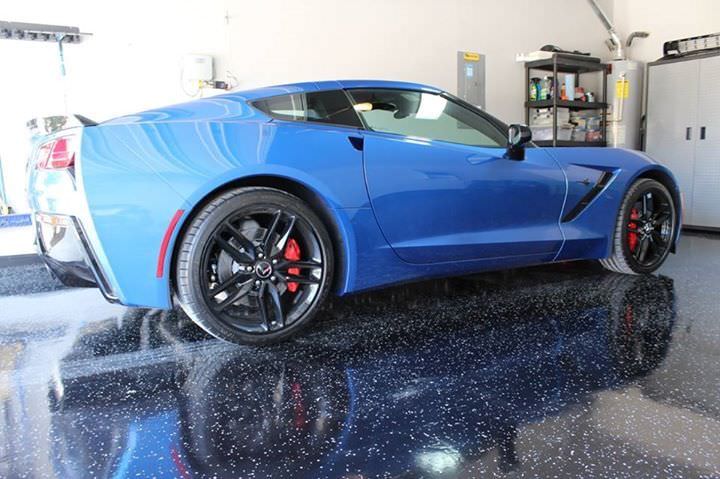 A vibrant blue sports car with sleek bodywork and black rims with red detailing is parked on a glossy epoxy garage floor with speckles. The reflective surface of the floor creates a mirror-like effect, highlighting the car's well-maintained condition. Visible in the background are garage storage units and equipment, suggesting a clean and organized space.