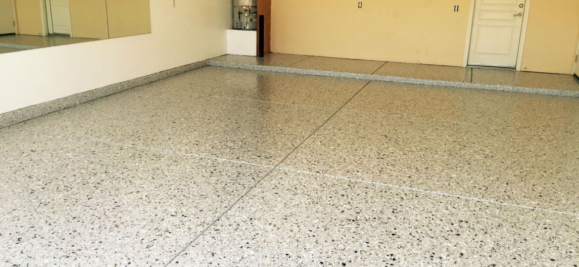 Spacious room with terrazzo coved flooring in neutral tones, featuring a large mirror on the left wall and a closed white door on the right wall. The room has a water heater and electrical panels on the left, and outlets and switches are visible on the pale yellow walls.