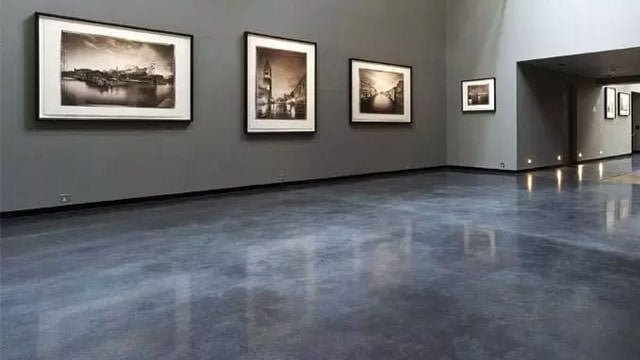 A spacious room with a polished concrete floor reflecting subtle light, creating a smooth and even surface. The gray floor complements the dark gray walls adorned with framed black and white photographs, suggesting an art gallery setting. The soft lighting accentuates the artworks and the clean, minimalist design of the space.