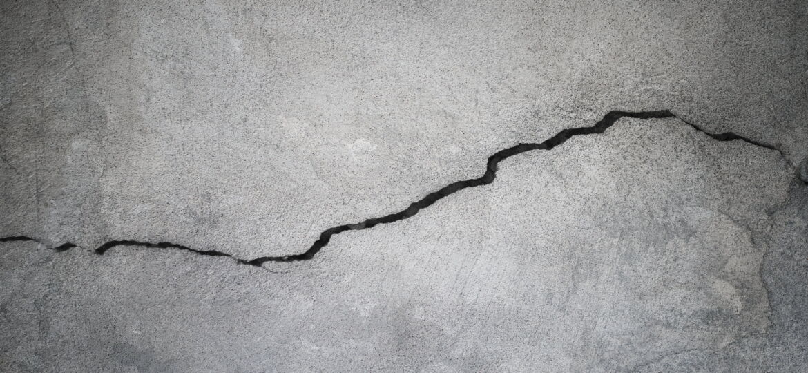 Close-up of a jagged crack running through a textured grey concrete wall, indicating structural damage or wear.
