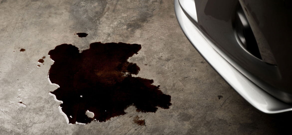 A dark stain on a concrete floor, shaped irregularly, suggesting a fluid leak near the front wheel of a vehicle, partially visible on the right. The texture of the floor and the shadow cast by the wheel suggest a garage or workshop environment. The exact nature of the fluid is unclear, possibly oil or another automotive fluid