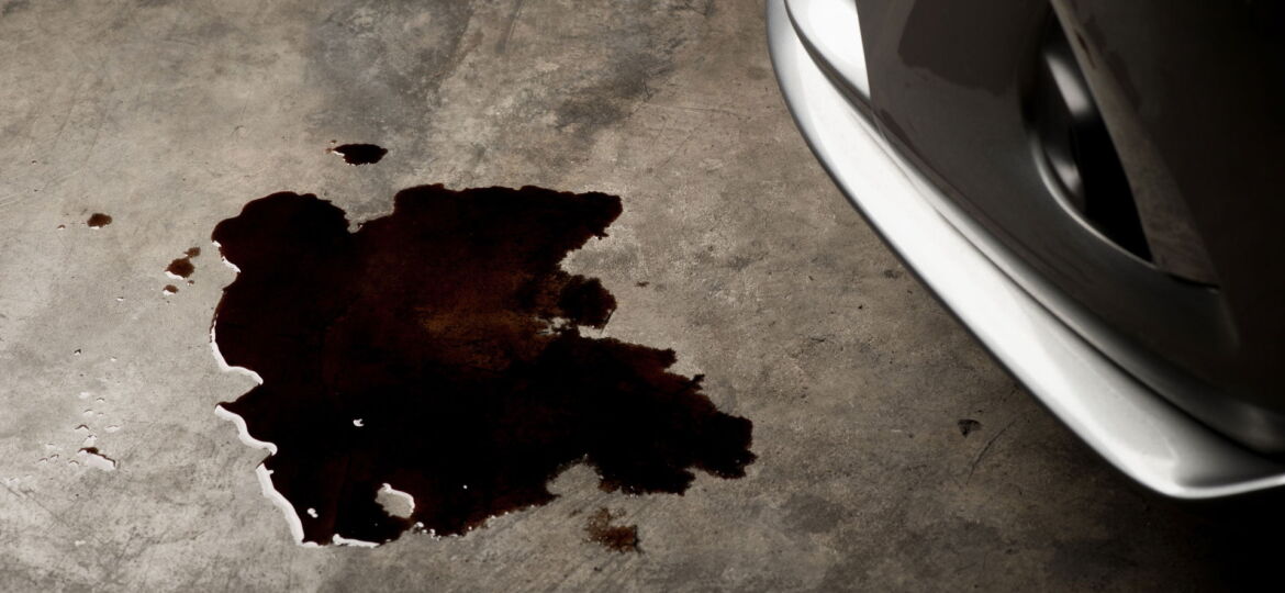 A dark oil stain on a concrete floor, shaped irregularly, suggesting a fluid leak near the front wheel of a vehicle, partially visible on the right. The texture of the floor and the shadow cast by the wheel suggest a garage or workshop environment. The exact nature of the fluid is unclear, possibly oil or another automotive fluid