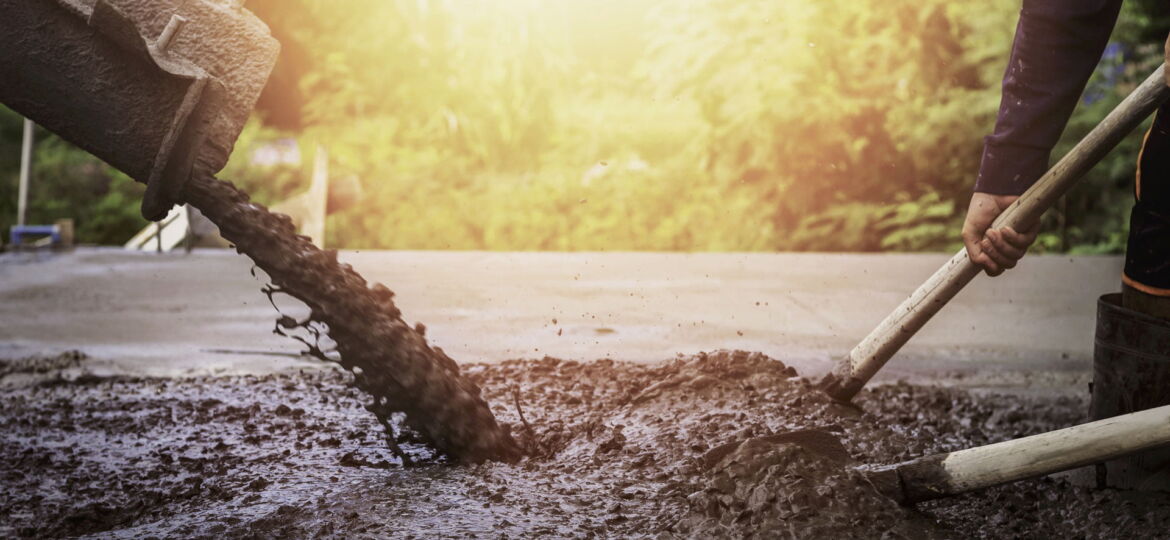 A construction worker in the process of pouring wet concrete from a wheelbarrow, using a shovel to spread the material for a new pavement or foundation. The scene is bathed in warm sunlight, indicating outdoor work possibly in a residential or park area.