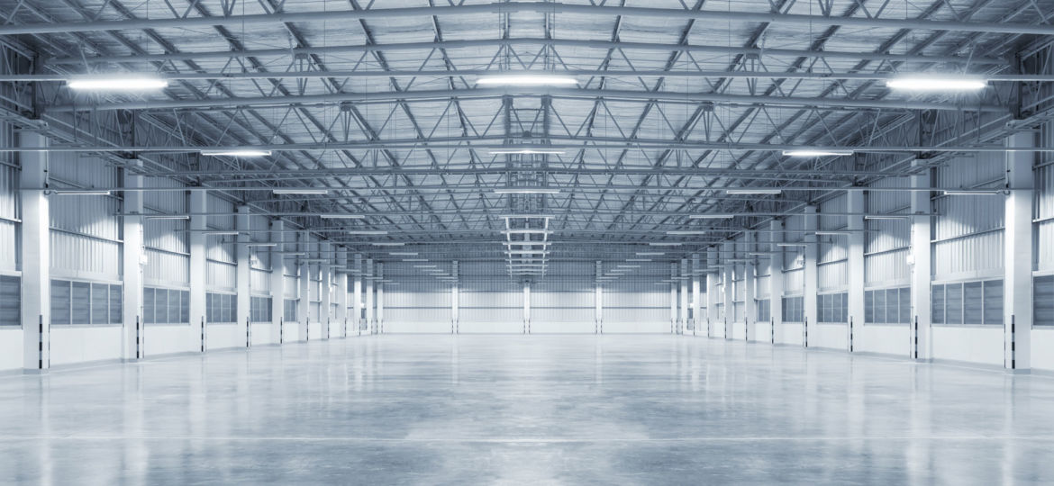 A vast, empty industrial warehouse with a high ceiling, supported by a network of metal trusses. The space is illuminated by a series of fluorescent lights and natural light streaming through the large windows on the side walls, reflecting on the polished concrete floor.