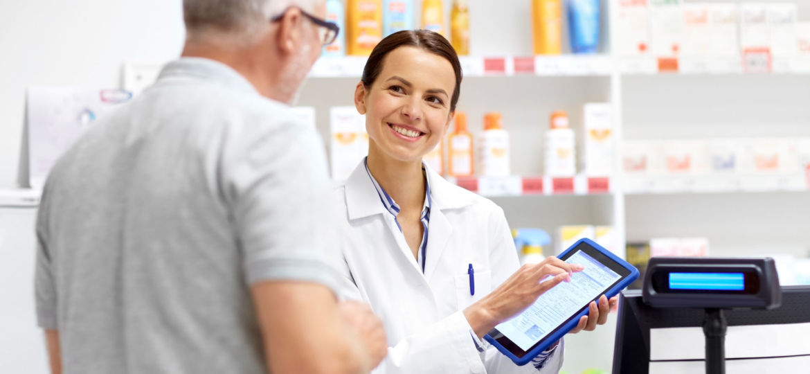 A smiling female pharmacist in a white coat is assisting an elderly male customer at a pharmacy. She is holding a digital tablet, possibly reviewing a prescription or patient information, with shelves of healthcare products in the background.