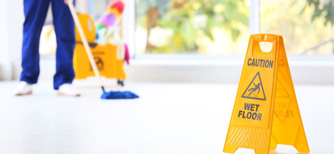 A vibrant yellow 'Caution Wet Floor' sign stands prominently in the foreground of a brightly lit room with a large window. In the background, an individual in blue attire is mopping the epoxy floor, and a janitorial cart equipped with cleaning supplies and colorful mops is visible, indicating active cleaning in progress.