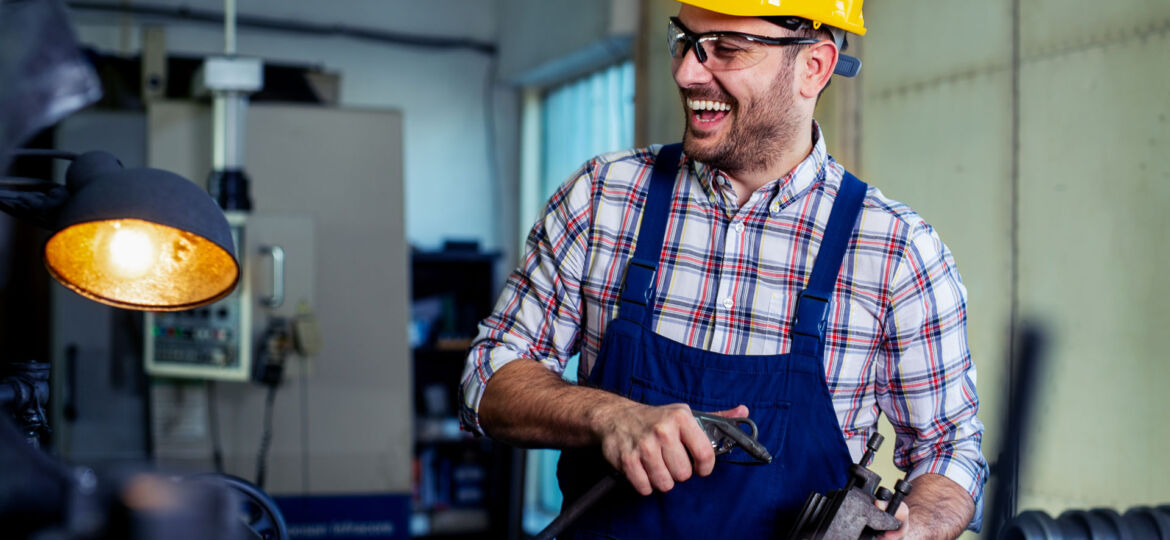 A cheerful worker wearing safety glasses and a yellow hard hat laughs while holding tools in a workshop. He's dressed in a plaid shirt with a blue overall, standing near machinery with industrial equipment in the background, under warm artificial lighting.