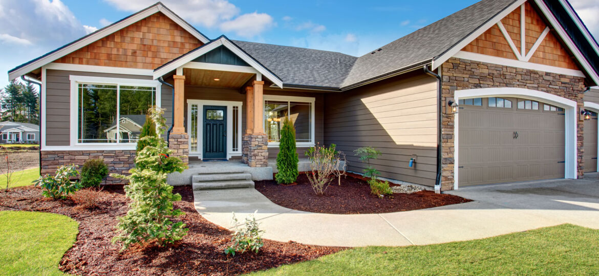 Modern single-story house with mixed siding featuring cedar shingles and stone accents, complemented by dark trim around windows and doors. A well-manicured lawn with mulched flower beds hosting shrubs and young trees leads to a concrete pathway towards the inviting front porch and a large garage door on the right.