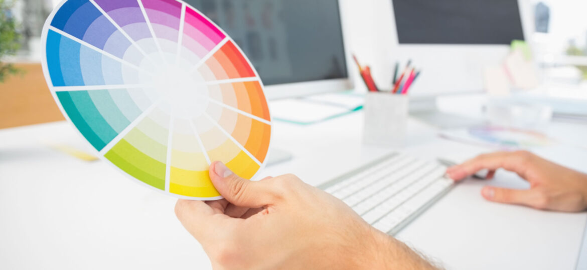 Close-up of a person's hands holding a color wheel with a spectrum of shades in front of a bright, modern workspace. The desk features a computer with a black screen, a keyboard, and a pencil holder filled with various writing tools, all under natural lighting.