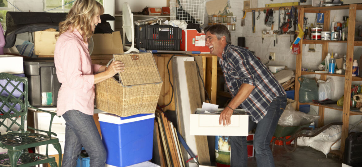 A man and woman organizing a cluttered garage, surrounded by various storage items like boxes, tools, and a wheelbarrow, indicating a cleaning or decluttering process in a domestic setting, possibly in spring time.