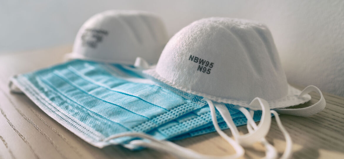 Two N95 respirator masks and a disposable blue surgical face mask resting on a wooden surface, indicative of health and safety measures in a medical or public health context.