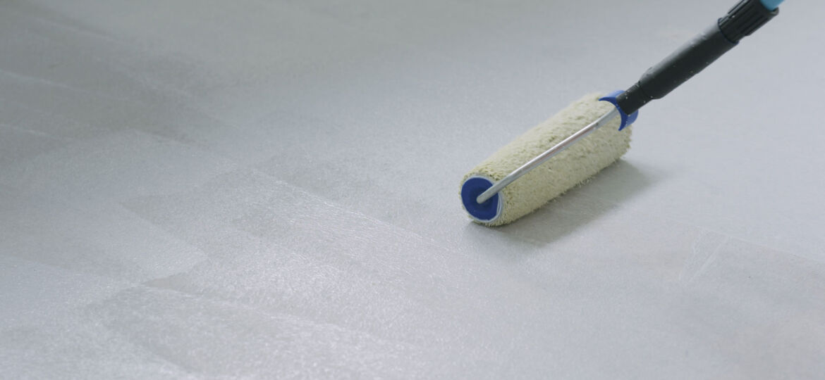 A paint roller with a blue handle being used to apply white epoxy paint on a concrete floor, leaving a wet paint trail and depicting a home improvement project in progress.