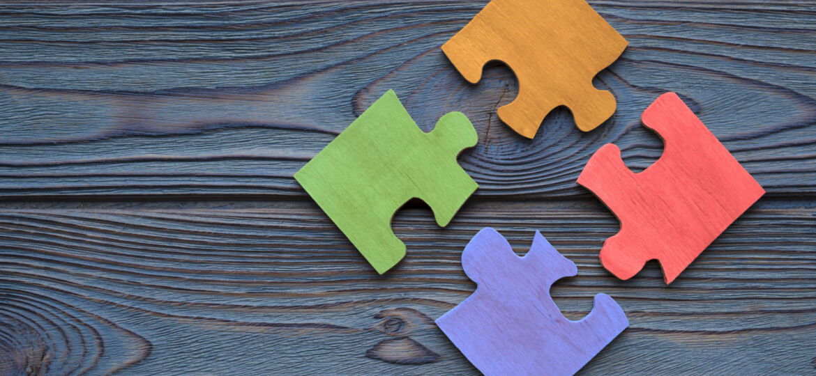 Four colorful jigsaw puzzle pieces in green, orange, red, and blue are scattered on a dark wood-grain surface, symbolizing problem-solving or the concept of fitting different elements together.
