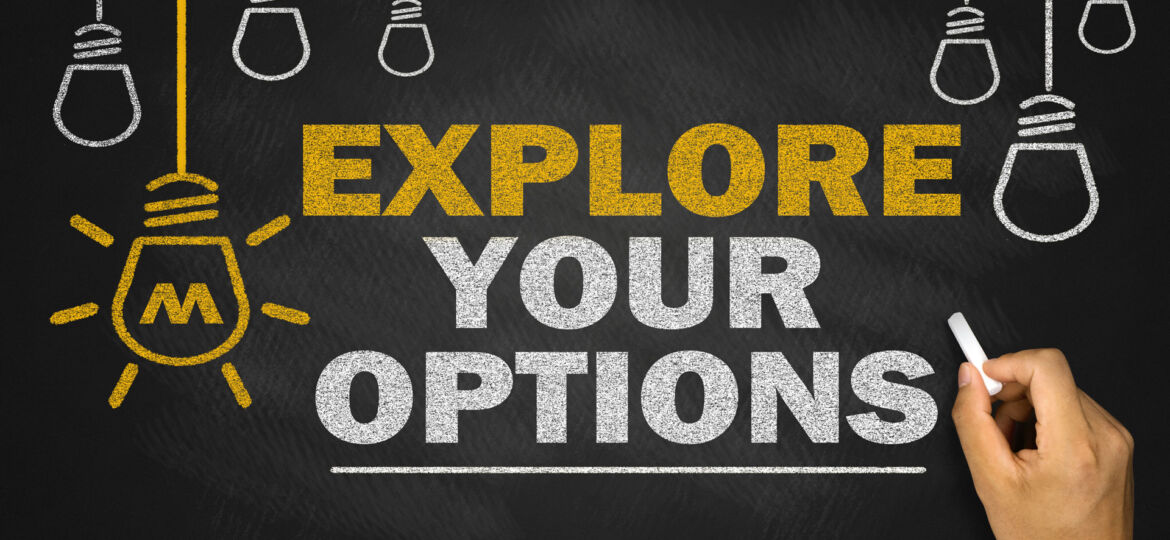 A hand writing on a black chalkboard with the phrase 'EXPLORE YOUR OPTIONS' in bold, yellow and white chalk. Above the text, drawn light bulbs hang from the top, with one bulb highlighted in yellow, symbolizing a bright idea or inspiration.