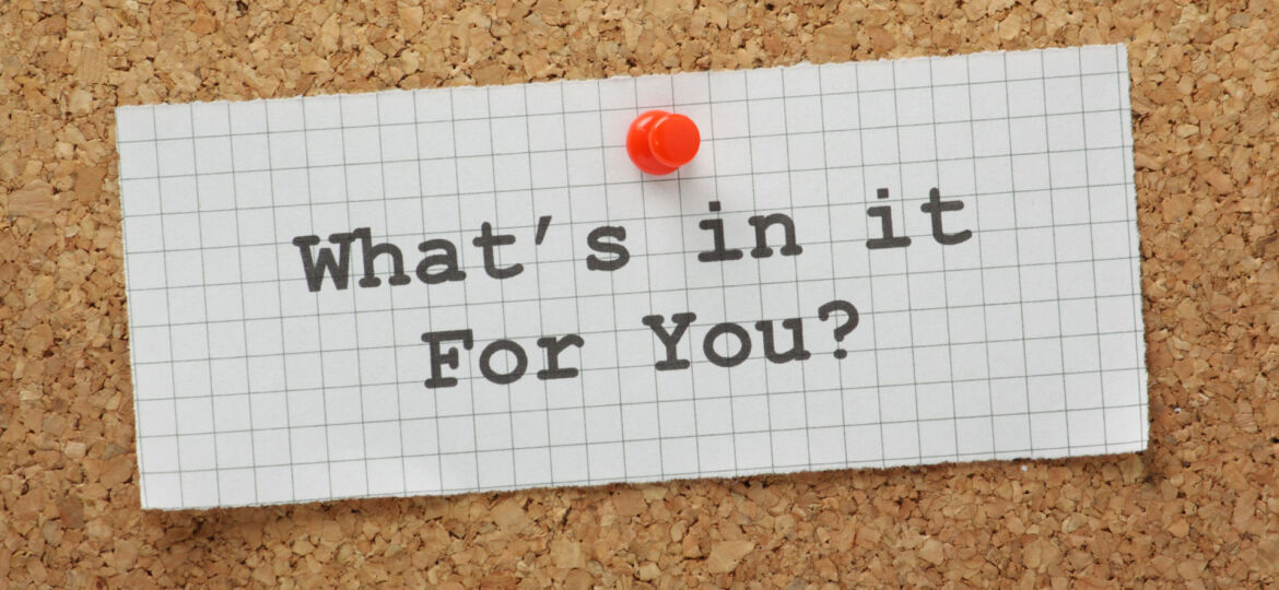 A piece of graph paper with the question 'What's in it For You?' printed on it, pinned to a cork bulletin board with a red pushpin.