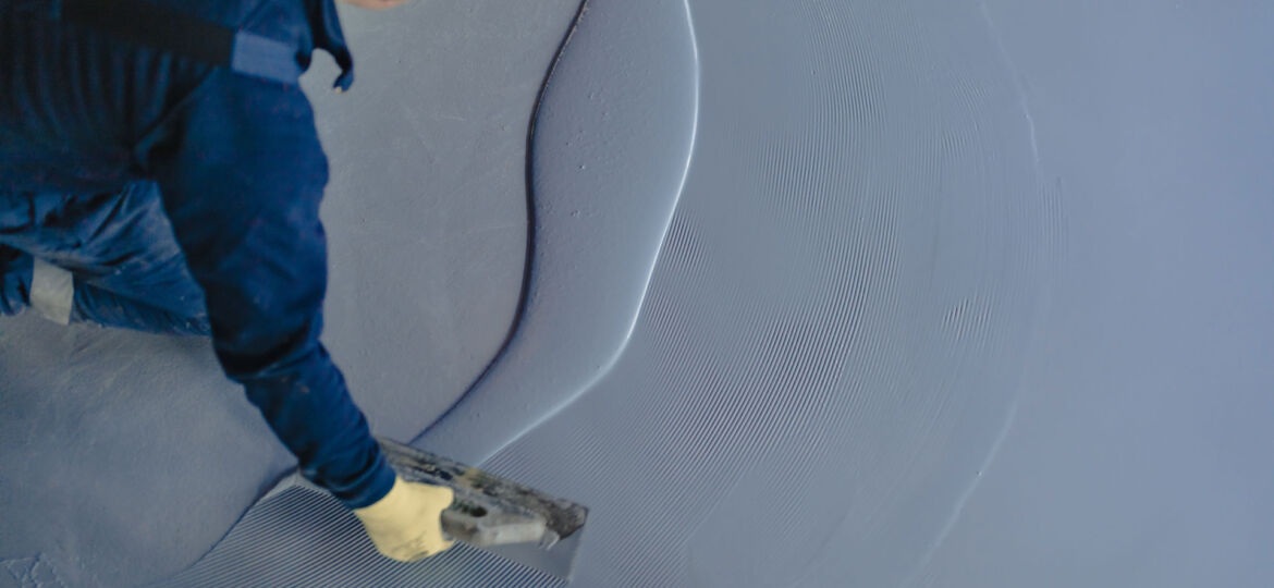 Overhead perspective of a worker applying gray self-leveling compound on a floor. The worker, wearing dark clothing and protective gloves, is smoothing the compound with a spreader, creating a seamless, even surface.