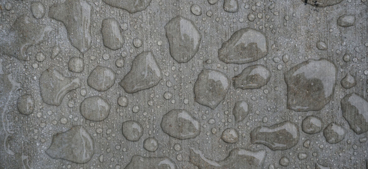 Textured surface of a concrete pavement embedded with various sizes of smooth pebbles and small stones, creating a speckled pattern typical of exposed aggregate finishes.
