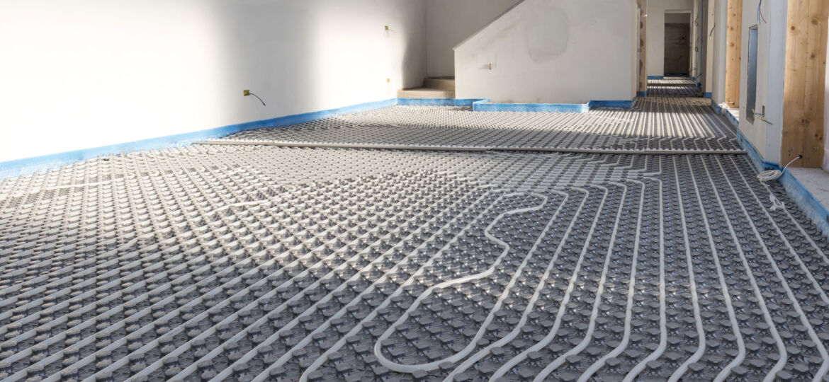 Interior view of an under-construction room with an underfloor heating installation. Visible are the serpentine arrangement of white heating pipes laid out on a gray insulating mat, with the perimeter edged by blue tape and walls prepared for plastering or painting.