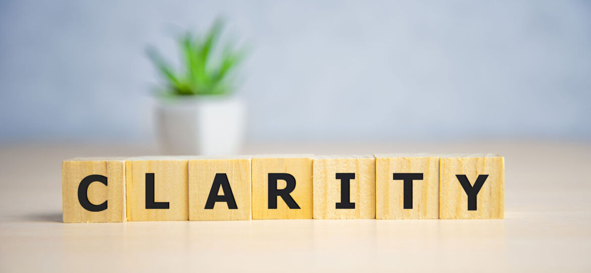 Wooden blocks with bold black letters spelling out the word 'CLARITY' on a desk, with a blurred potted plant in the background, conveying the concept of clear thinking or transparency in a calm and serene setting.