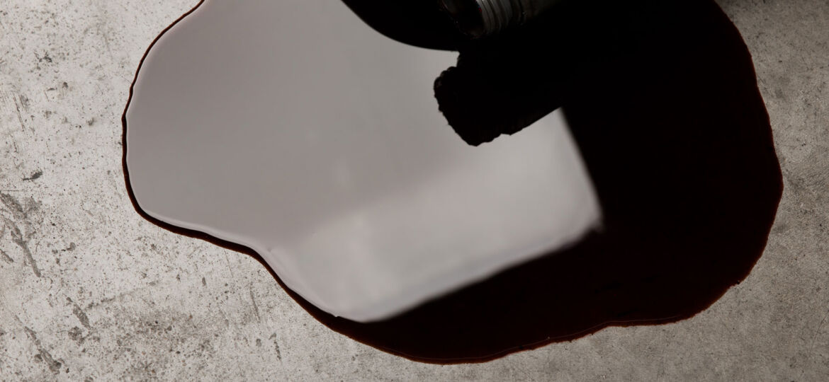 A close-up of a dark viscous liquid, possibly oil, spilling from an overturned container onto a concrete surface, illustrating an accidental spill with a focus on the fluid's glossy texture and the contrast against the rough ground.