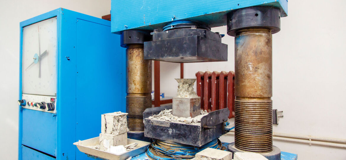 A heavy-duty hydraulic compression testing machine in a laboratory setting, with a blue frame and pressure gauge, crushing a concrete cube to determine its compressive strength. Pieces of fractured concrete are visible around the machine, indicating a completed strength test.