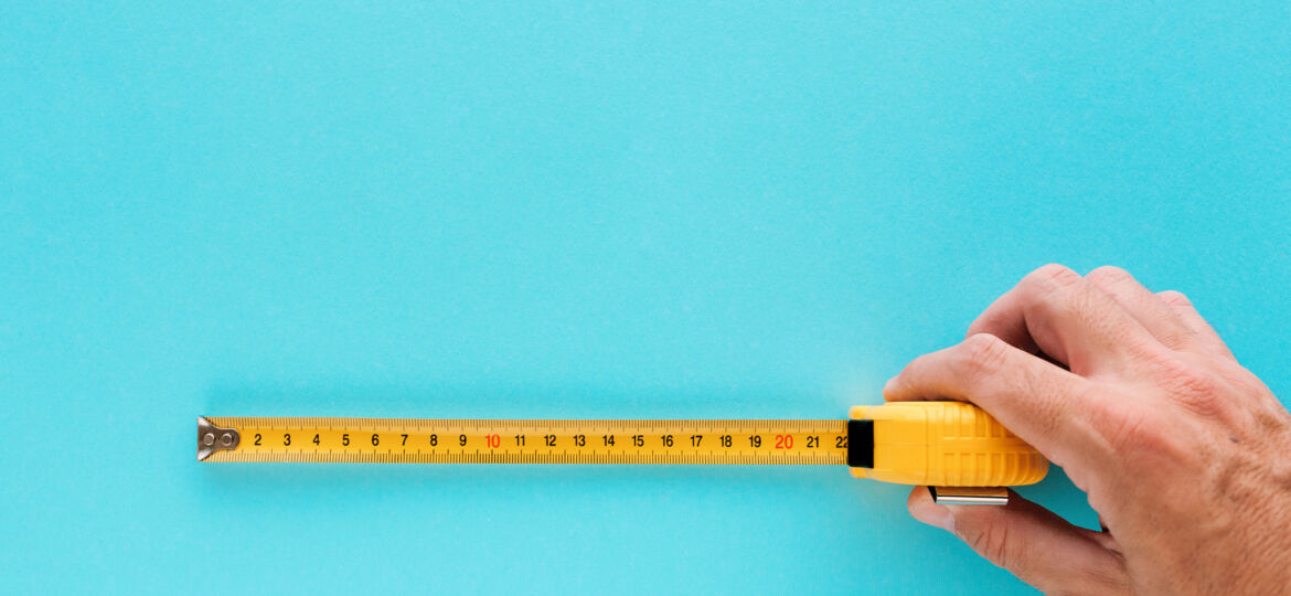 A hand holding a yellow tape measure extended across a solid turquoise background. The tape measure shows centimeters and is extended to the 22-centimeter mark, indicating the measurement being taken.