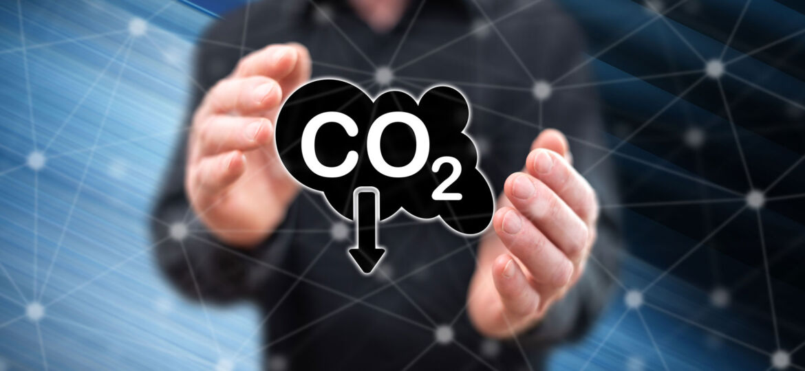 A person's hands are framed as if holding an invisible object, with a digital illustration of a CO2 cloud icon with an arrow pointing downwards, symbolizing carbon dioxide reduction. The background is a dynamic blue with a network grid, suggesting a high-tech or scientific context.