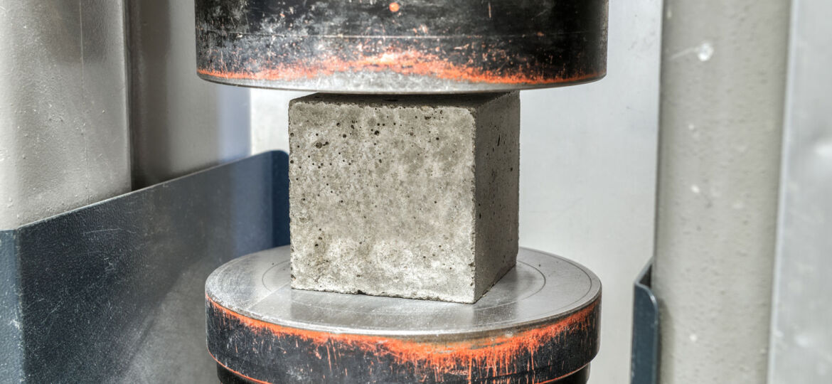 A close-up of a compression test in progress, featuring a concrete cube sample placed between the plates of a testing machine. The upper plate is in contact with the cube, applying pressure to assess the material's strength and durability, a common quality control measure in construction.