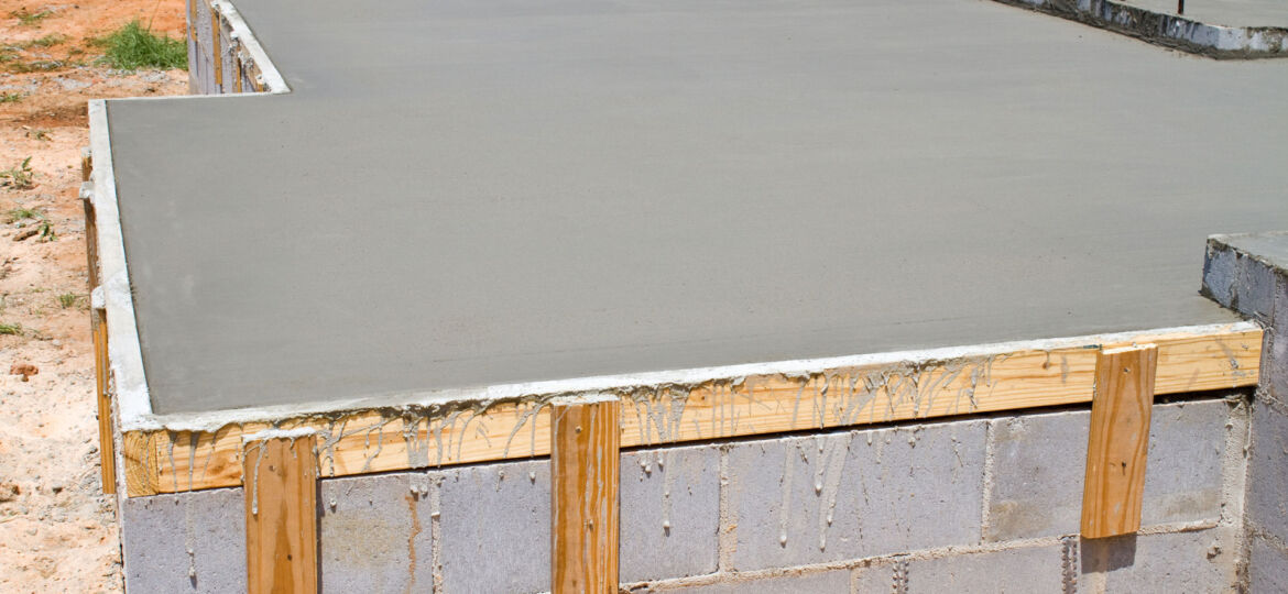 Freshly poured concrete slab with wooden formwork still attached at the edges. The slab is atop a cinder block foundation, with metal rebar protruding in preparation for additional structural support. This image captures an early stage in the construction process of a building's foundation.