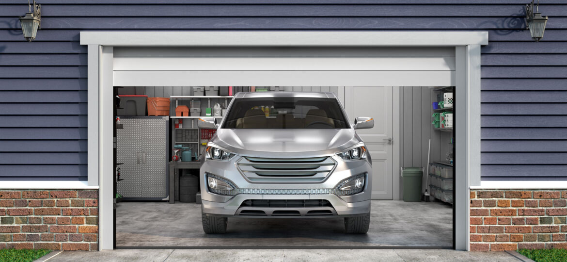 A silver SUV is parked inside a tidy residential garage, with the garage door open. The garage has organized shelves on the sides, storing various items like containers, tools, and household supplies. The exterior of the garage features navy blue siding and a brick base, complementing the clean and organized appearance of the home.