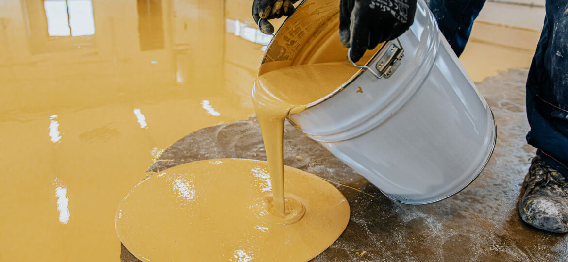 A person in work attire and gloves is pouring a thick, yellow epoxy paint liquid from a large bucket onto a concrete floor, with the liquid spreading out on the surface. The setting suggests an industrial or construction site where the floor is being coated or treated with a specific substance.