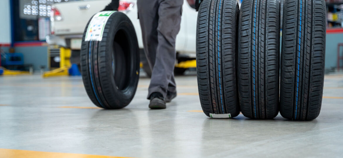 In a commercial automotive service center, a set of four new black tires are prominently displayed in the foreground. In the background, a mechanic in a green and black uniform is walking away, partially visible, with another car raised on a lift, indicating an active work environment focused on vehicle maintenance.