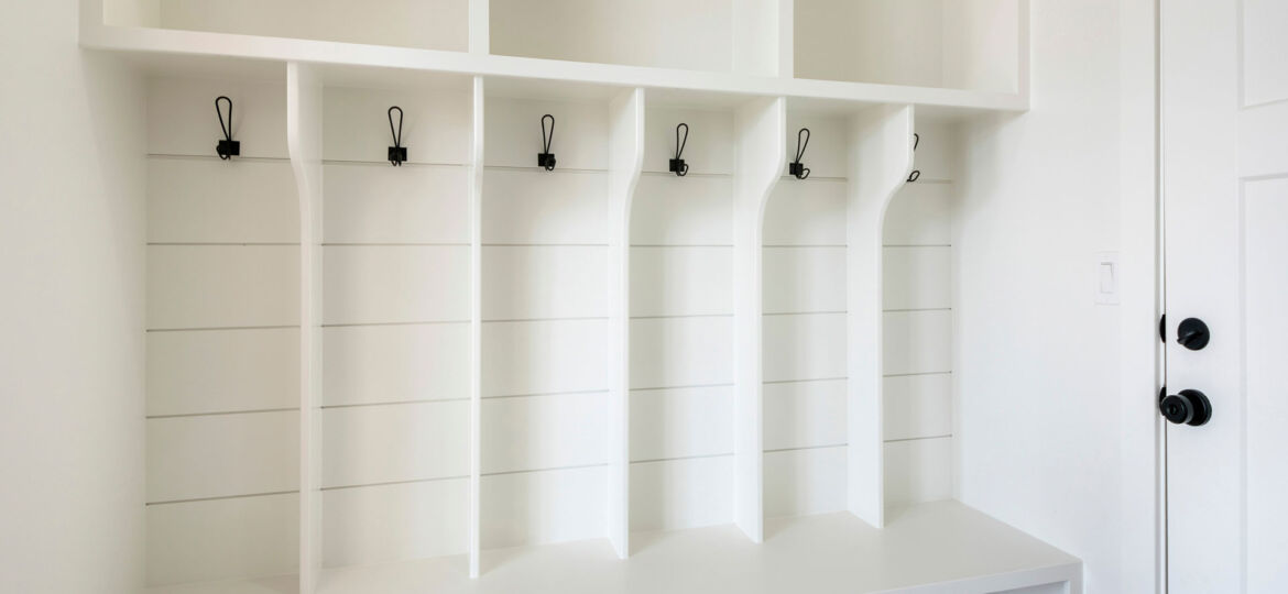 A modern, clean white mudroom with built-in shelving and bench. Five black metal hooks are evenly spaced along the upper section for hanging items, and the design features simple, straight lines. To the right, a closed door with a black doorknob complements the minimalist aesthetic of the space.