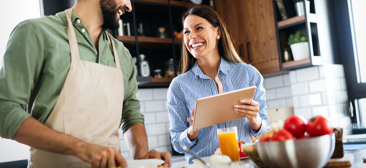 A man in an apron and a woman holding a tablet are sharing a joyful moment in a kitchen. The man is chopping vegetables on a cutting board, while the woman, with a broad smile, appears to be looking at a recipe or a cooking tutorial. The kitchen is modern and bright, with fresh tomatoes and a bowl of fruit on the countertop, suggesting a healthy cooking activity.