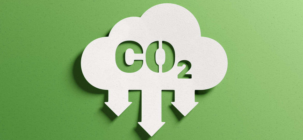 A white cut-out of a cloud with the chemical symbol 'CO2' in the center and three arrows pointing downwards, placed against a bright green background. This imagery symbolizes the concept of carbon dioxide reduction or carbon footprint awareness.