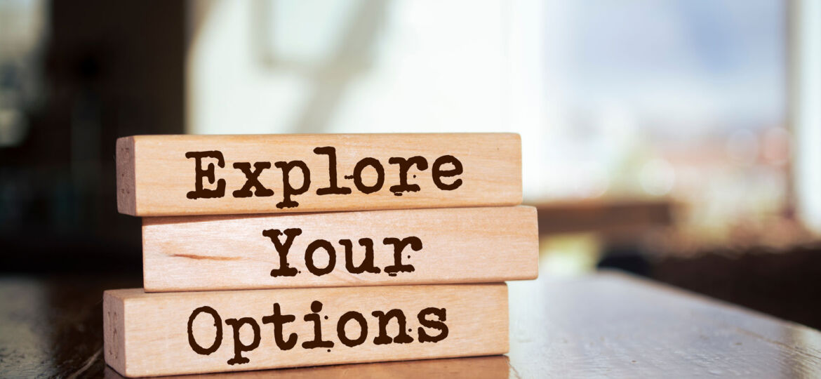 Three wooden blocks stacked on top of each other on a shiny wooden surface, with the words 'Explore Your Options' burned into the wood in capital letters. The background is softly focused, showing light streaming in through a window, suggesting a bright, contemplative space.