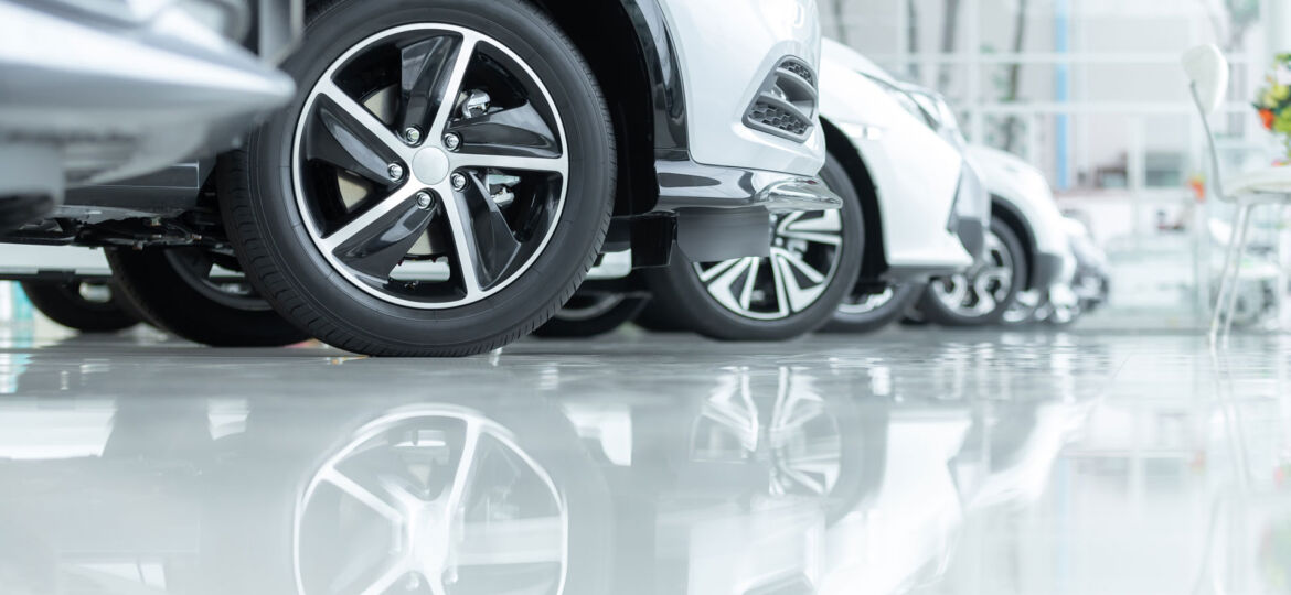 A row of new cars on display at a dealership, with a focus on the shiny alloy wheels of the nearest car. The vehicles are parked on a glossy floor, which reflects the wheels and lower car bodies, enhancing the pristine condition and polished appearance of the showroom.