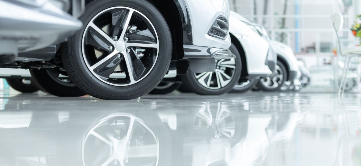 A row of new cars on display at a dealership, with a focus on the shiny alloy wheels of the nearest car. The vehicles are parked on a glossy epoxy floor, which reflects the wheels and lower car bodies, enhancing the pristine condition and polished appearance of the showroom.