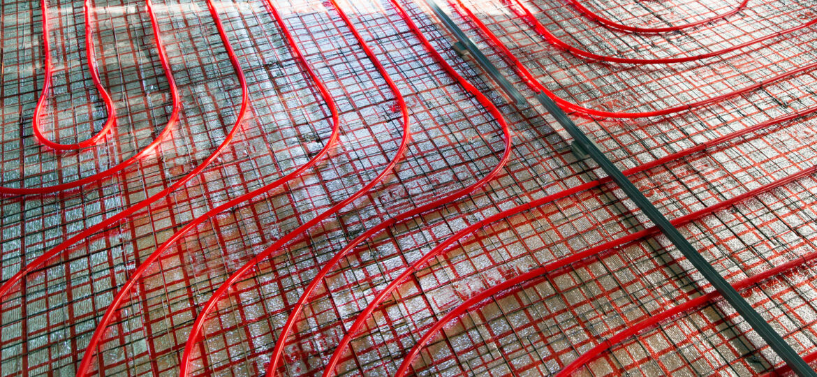 Close-up view of a red underfloor heating installation. The system comprises a network of red pipes arranged in a serpentine pattern over a mesh reinforcement layer on a concrete floor, indicating the preparatory phase of underfloor heating before the final flooring is laid.