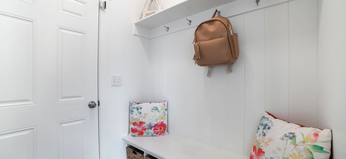 A neatly organized mudroom corner with a white built-in bench seat adorned with floral cushions. Above the bench, a tan backpack hangs from one of the hooks on the white paneled wall, and wicker storage baskets are tucked underneath the bench.