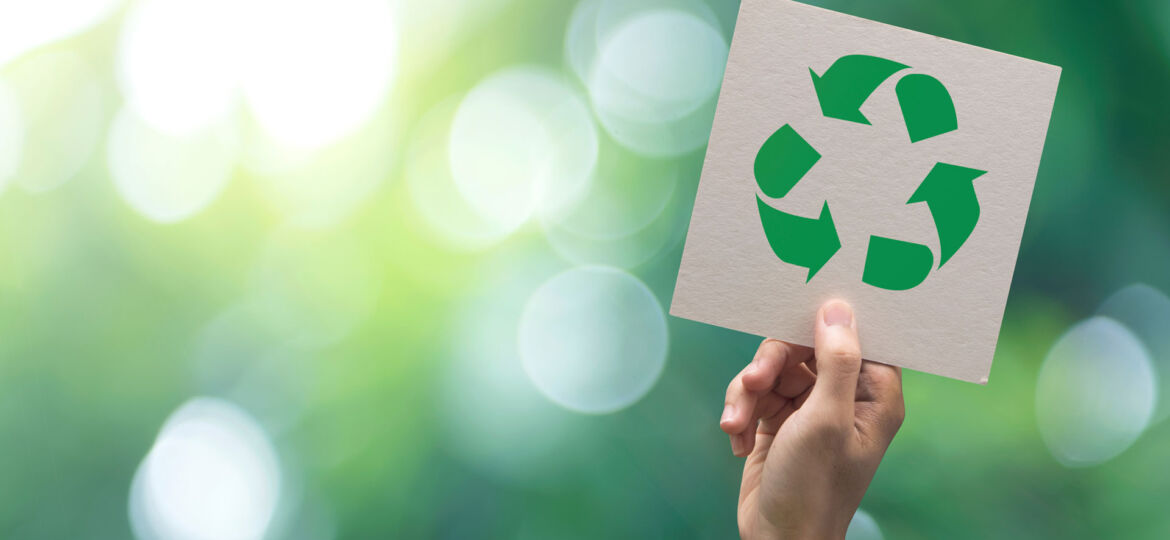 A hand holding up a card with the recycling symbol cut out, against a bright, bokeh background of green foliage, symbolizing environmental awareness and the importance of recycling for nature conservation.