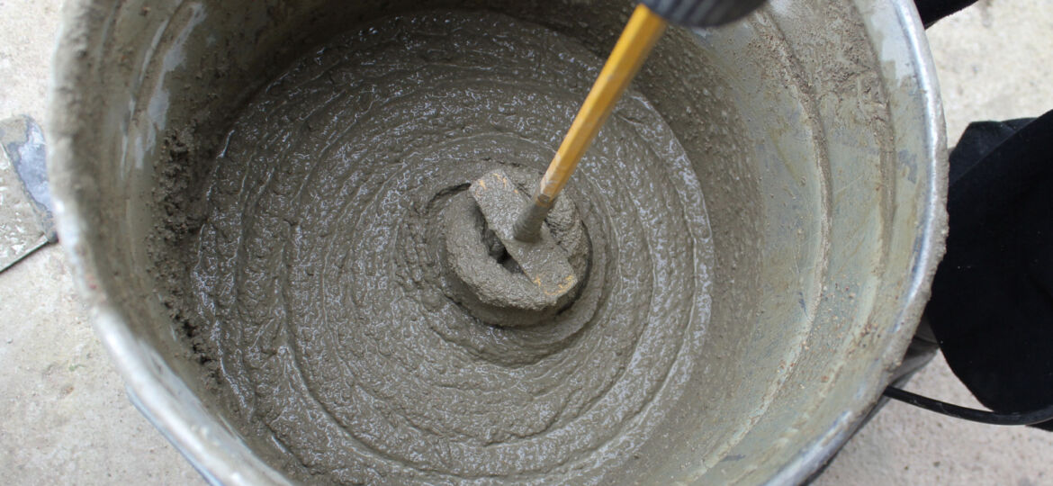 Overhead view of a bucket of freshly mixed concrete with a spiral mixing attachment on a power drill immersed in it, capturing the process of preparing material for construction work.