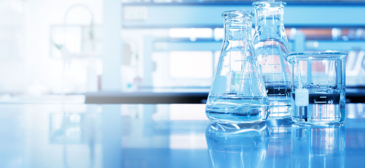 A bright, modern laboratory setting with a close-up of clear glassware, including Erlenmeyer flasks and a beaker filled with liquid, reflecting on the shiny surface of a lab bench with a soft-focus background.