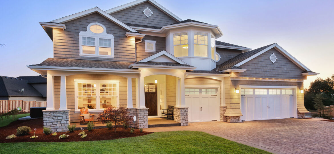 Twilight view of a large two-story suburban home with a stone and siding exterior, featuring a covered porch with a rocking chair and a double garage. The home's interior and exterior lights are on, creating a warm and inviting atmosphere.
