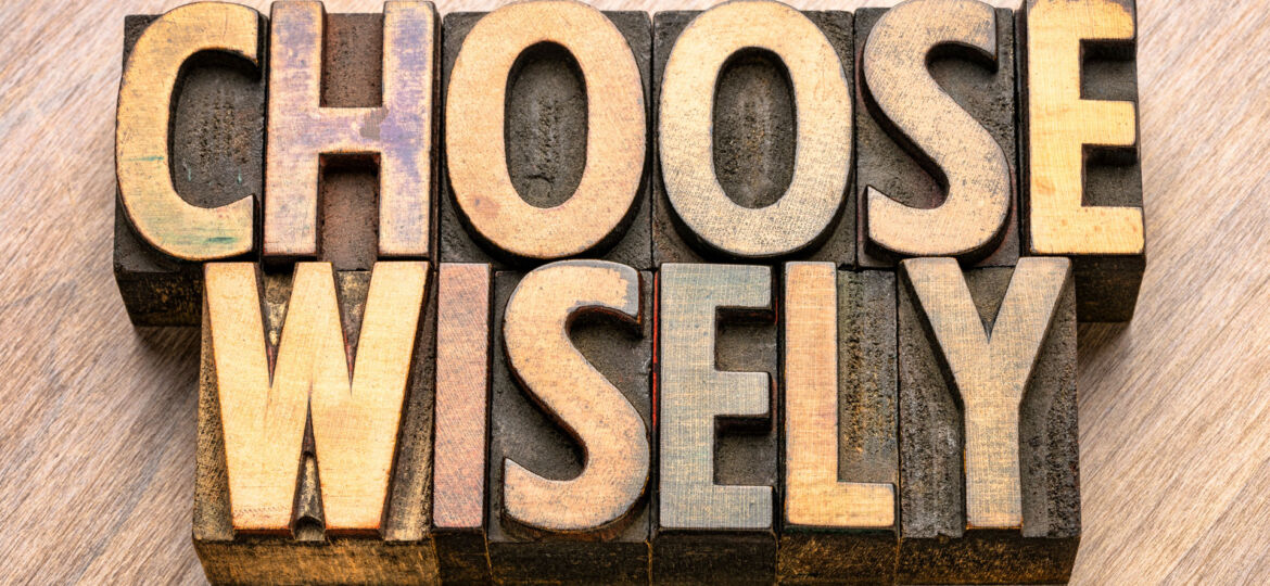 The phrase 'CHOOSE WISELY' spelled out in bold, vintage wooden letterpress printing blocks on a wood-grain background, highlighting the importance of thoughtful decision-making.