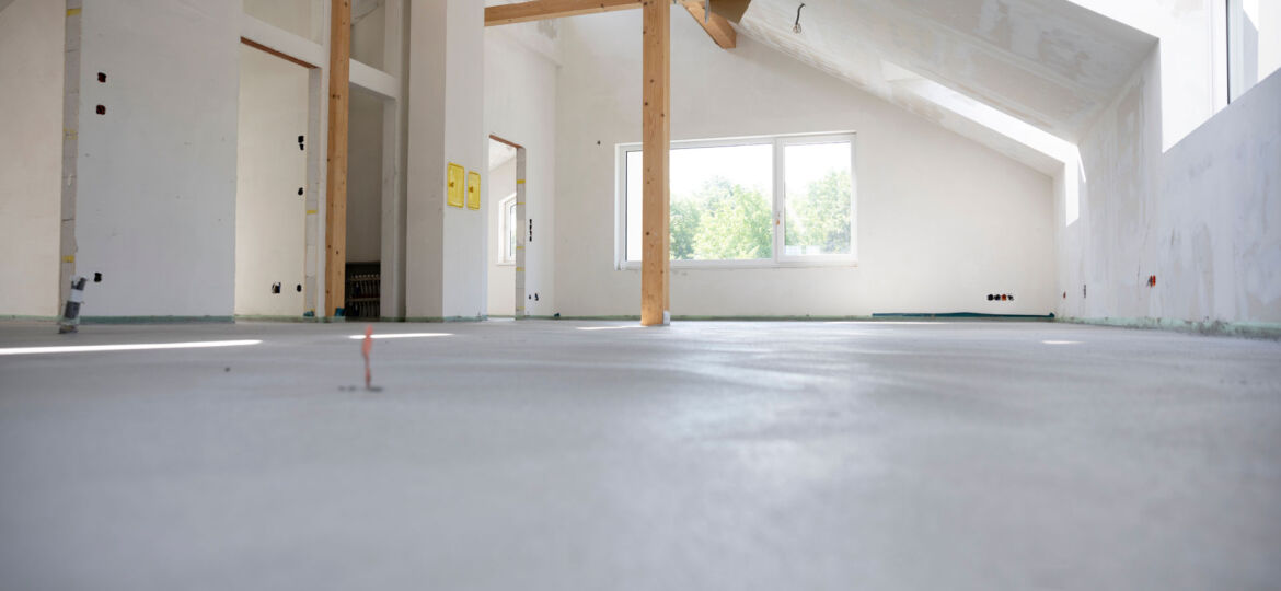 Interior of a room under construction with unfinished walls and exposed wooden beams. The space is well-lit by natural light from windows and the concrete floor is smooth and bare, with various construction marks. The walls are primed for painting or further finishing, depicting an early stage of renovation.